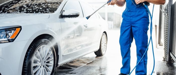 Professional washer in blue uniform washing luxury car with water gun on an open air car wash. Close up photo.